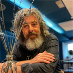Photograph of Dave smiling, he is leaning over with his hand on his arm. He has long grey hair and a long beard