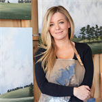 image of debbie smiling, she has long blonde hair and is wearing an apron with paint splatters, she is sitting in front of two painting portaits